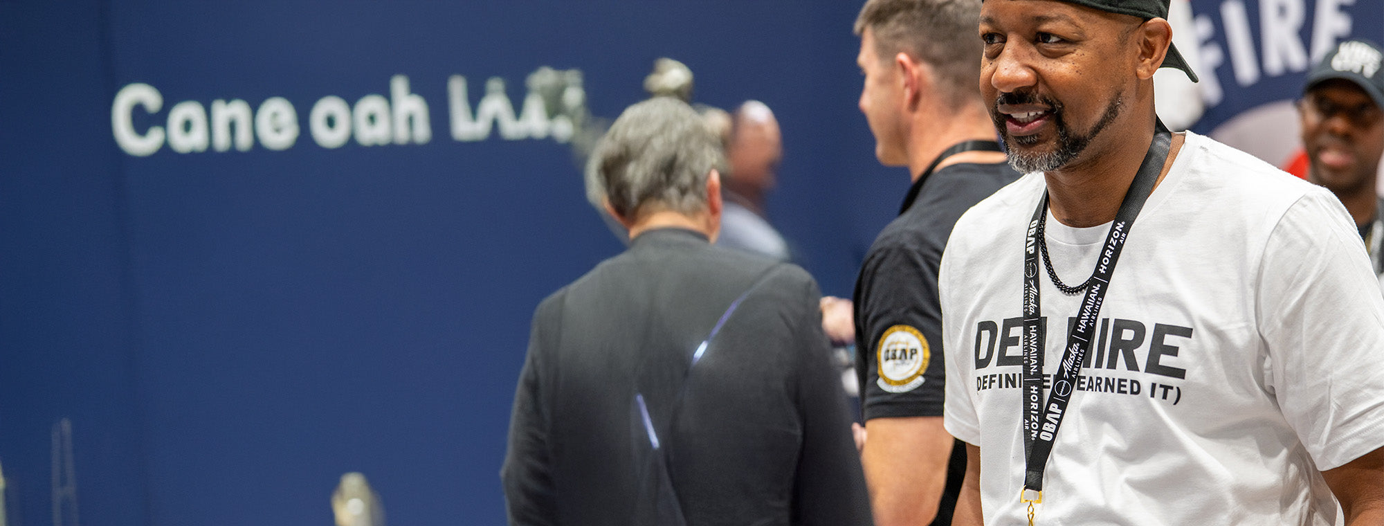 Man wearing a white shirt with 'DEI HIRE' on it, standing in front of a blue wall with text.